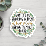 Sticker with text 'Just a girl standing in front of her plants asking them not to die' on a wooden surface with plants.