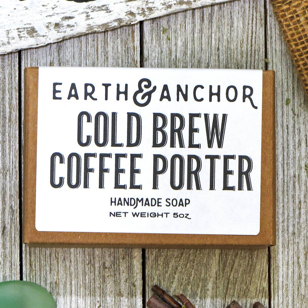 A packaged bar of Cold Brew Coffee Porter handcrafted vegan soap by E&A, displayed on a wooden surface with decorative items around it.
