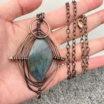 Hand holding a copper wire-wrapped pendant with a blue stone on a neutral background