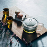 Clear glass teapot with green liquid, two glasses of tea, macarons, and lavender on a wooden board.
