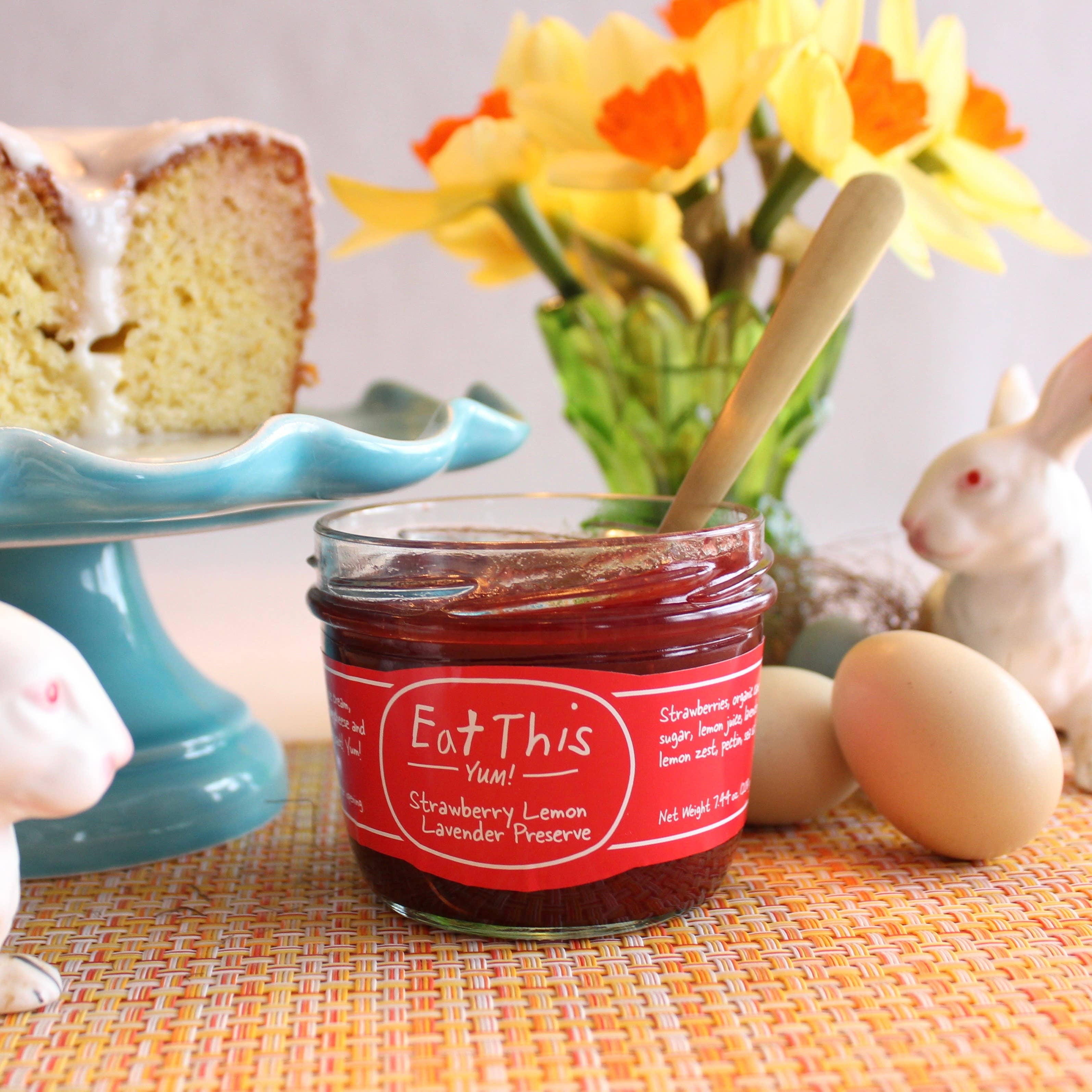 Jar of strawberry lemon lavender preserve with a spoon, cake, and flowers on a table.