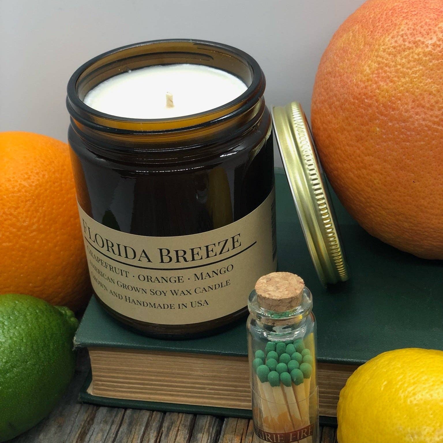 Candle labeled 'Florida Breeze' with oranges, a book, and matches on a wooden surface.