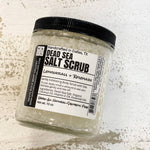 A clear plastic jar containing a white or light gray dead sea salt scrub, labeled with information about the product.