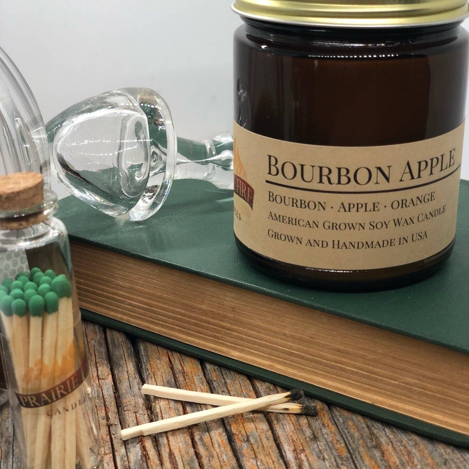Jar of Bourbon Apple candle with matches and a book on a wooden surface