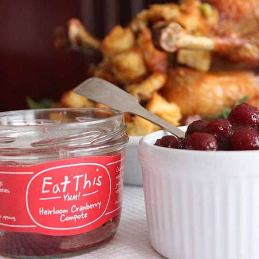 Jar of cranberry compote with a label, bowl of cranberries, and roasted turkey in the background.