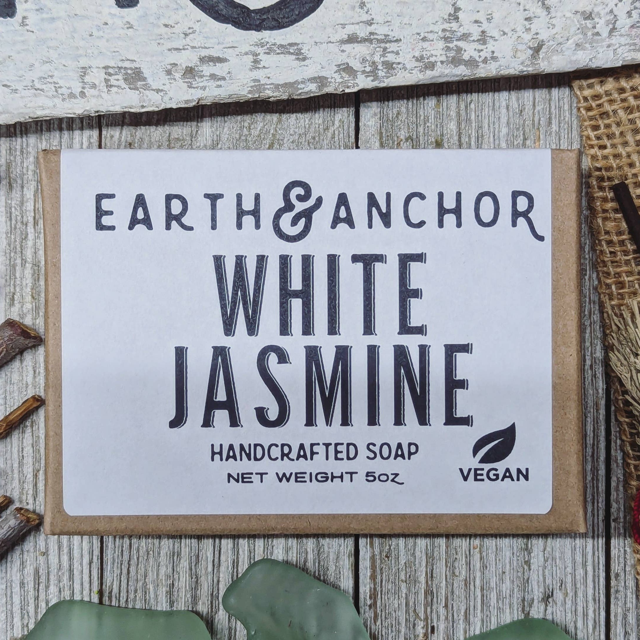 A packaged bar of handcrafted soap labeled 'White Jasmine' from Earth & Anchor, displayed on a wooden surface with decorative elements around it.