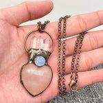 A handmade pendant necklace featuring a heart-shaped pale pink stone, three clear crystal points, a round blue stone, and an antique bronze metal frame, held in a person's hand.