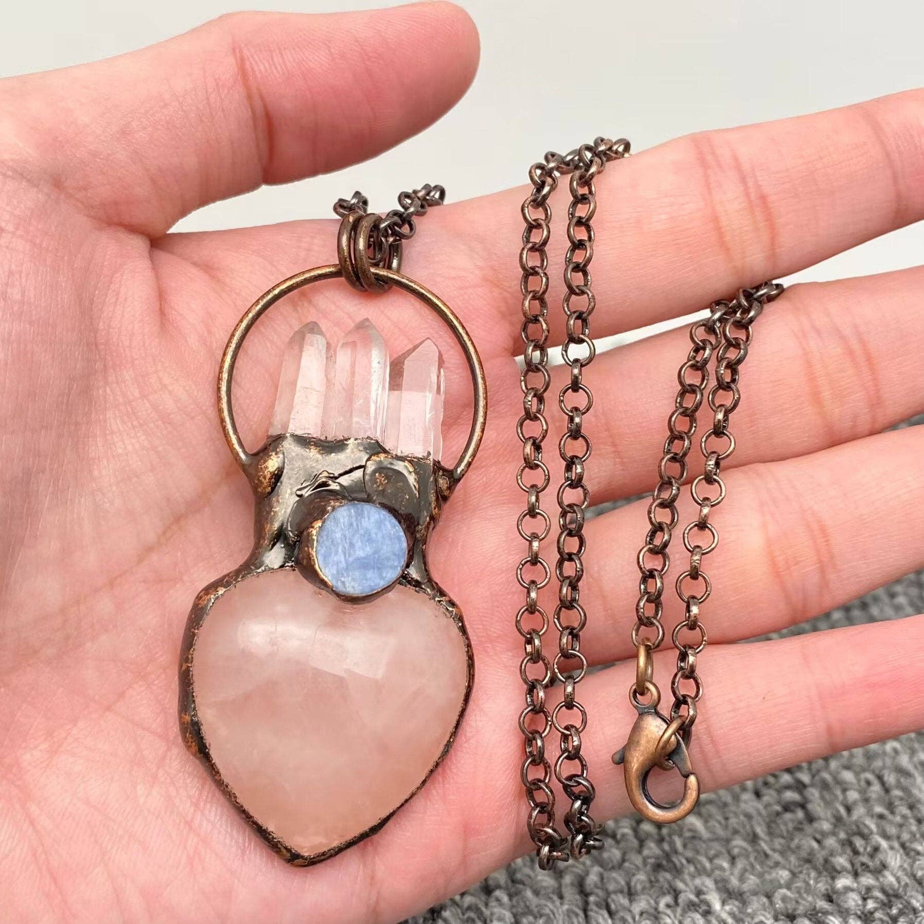 A handmade pendant necklace featuring a heart-shaped pale pink stone, three clear crystal points, a round blue stone, and an antique bronze metal frame, held in a person's hand.
