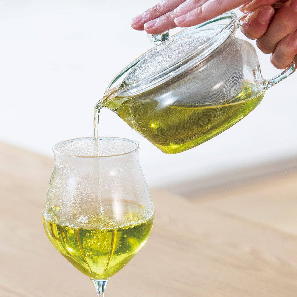 Hand pouring green liquid from a glass teapot into a glass on a blurred background