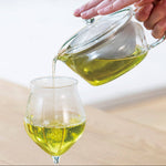 Hand pouring green liquid from a glass teapot into a glass on a blurred background