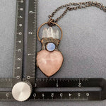A handmade pendant necklace featuring a pink rose quartz heart, a clear quartz crystal cluster, a small blue moonstone, and an antique bronze ring and chain, shown with metal rulers for scale on a textured gray background.