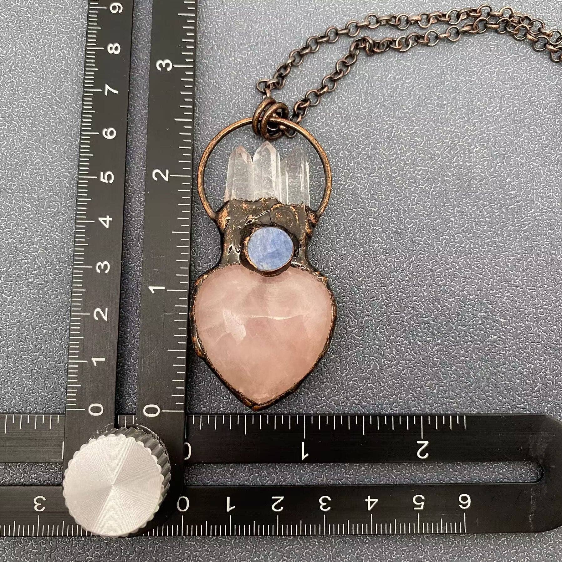 A handmade pendant necklace featuring a pink rose quartz heart, a clear quartz crystal cluster, a small blue moonstone, and an antique bronze ring and chain, shown with metal rulers for scale on a textured gray background.