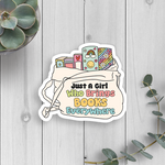 Sticker with 'Just A Girl Who Brings Books Everywhere' text on a wooden surface with plants.