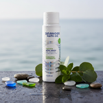 A bottle of Seaweed Bath Co Hydrate Body Wash stands on a wet dark surface with decorative stones, eucalyptus leaves, and mint leaves, against a blurred ocean background.
