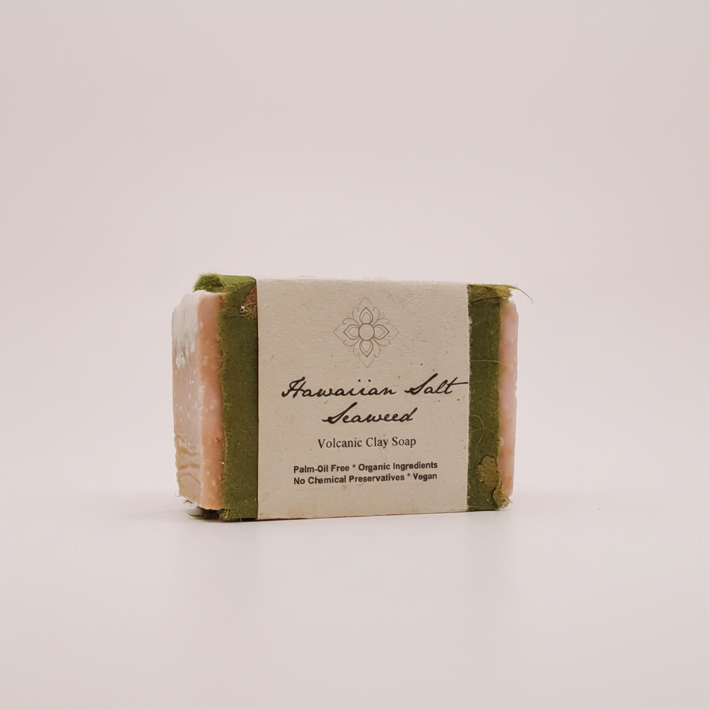 A wrapped bar of soap standing upright on a plain light background. The soap has green and pale pink sides with a cream paper band label reading 'Hawaiian Salt Kavaed Volcanic Clay Soap.