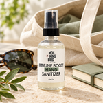 Mockingbird Apothecary Immune Boost Hand Sanitizer beside a canvas tote, sunglasses, sage-green notebook and leafy sprig on a stone surface.