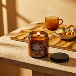 Lit Mulled Cider candle on travertine with an amber cider cup, ginger, cinnamon leaves and orange peel