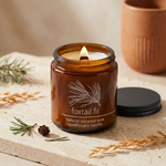 Lit Foxtail Fir amber apothecary candle with a ridged black lid on a warm stone tabletop with evergreen accents