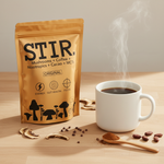 Stir Mushroom Coffee gold pouch on wood surface beside a white mug, wooden spoon with powder, dried mushrooms and cacao nibs scattered nearby