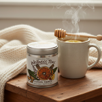 Tonsil Tamer Herbal Tisane tin beside tall stoneware mug of steaming golden tea with honey dipper and lemon on bedside table