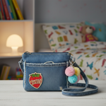 Small strawberry embroidered patch on worn tattered denim purse with pompom keychain on white table in cozy child's room