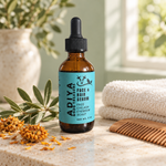 Adiya God's Treasure hair serum beside ivory towels, a wooden comb, olive leaves and dried flowers on a sunlit stone vanity.