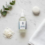 Unscented Magnesium Oil Spray – flat lay on white marble with magnesium flakes, white stone, eucalyptus and white linen