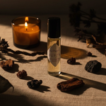 Adia Depth Fragrance Oil styled with amber candle, dark wood chips, and dried botanicals on cream linen