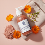 Pink Clay & Calendula Intense Hydrate Cleanser flat lay on blush surface with pink clay powder and calendula flowers