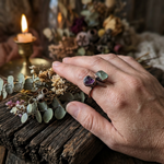 Creative lifestyle photo of the raw purple and green fluorite wrap-around ring worn on a hand over weathered wood with dried botanicals and candlelight