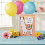 Funny llama birthday card propped on table with colorful balloons, wrapped gift and confetti