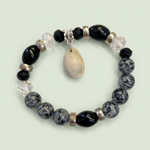 Black and gray beaded stretch bracelet with cream shell charm on a pale sage background