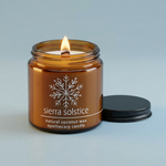 Hemlock Park Sierra Solstice 4 oz amber candle with a lit wood wick and ridged black lid on a soft slate-blue background
