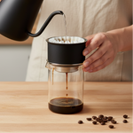 Fellow Stagg X Short pour over set assembled mid-pour with gooseneck kettle, coffee dripping into glass carafe on wood surface