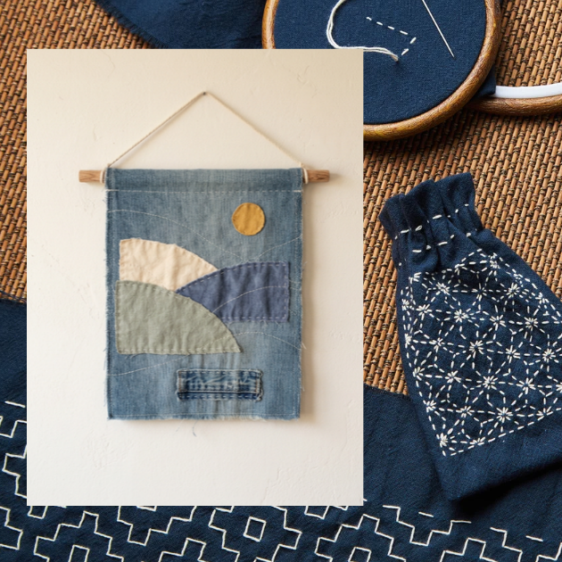 Denim wall hanging with patchwork hills and a golden sun, surrounded by indigo fabric with white sashiko stitching and an embroidery hoop.