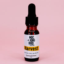 A small dark glass bottle with a dropper filled with Harvest Essential Oil Blend, labeled with white and orange text against a pink background.